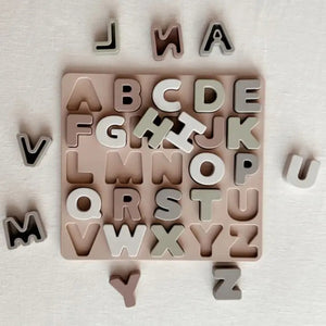 The ABC SOFT SILICONE PUZZLE from ALI + OLI sits on a light-colored surface, featuring cut-out slots for the entire alphabet. Some letter pieces are placed in their corresponding slots, while others are scattered around the board. This educational toy, with letters in various shades of beige, gray, and brown, promotes hand-eye coordination.