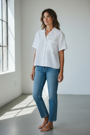 A woman stands barefoot in a bright room, smiling gently with her hands relaxed at her sides. She is wearing the LLEA BLUE - CASEY Short Sleeve Shirt in white from LLEA BLUE paired with blue jeans, as light pours in through large windows on the left.