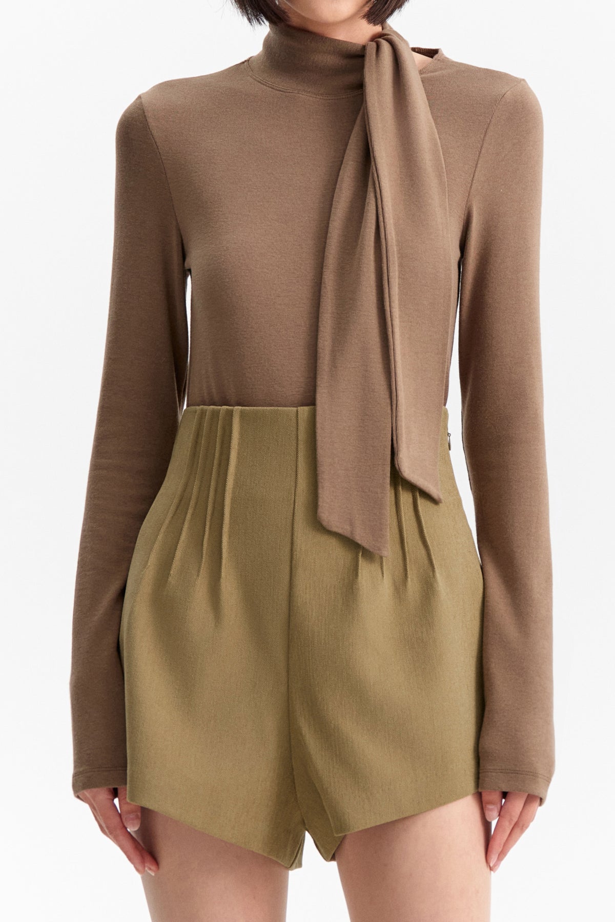 The ELEH - THE SCALA TIE-NECK TOP by ELEH is a light brown, long-sleeved minimalist blouse featuring an asymmetrical attached scarf draped across the front, shown on a hanger against a plain white background.