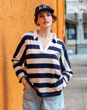 Wearing the MERSEA Amelia Rugby Tee in Navy Ecru Stripe, a woman pairs it with light denim shorts and a navy KC cap as she stands against an orange textured wall on a city sidewalk, gazing slightly to the side.