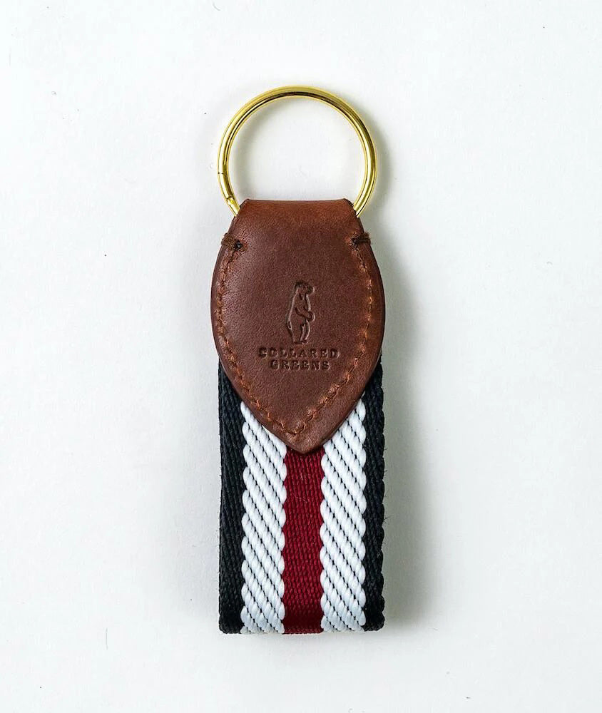 The BIRD DOG BAY COLLARED GREENS Key Chain features a gold ring, brown leather top with embossed logo, and a maroon, black, and white striped woven loop—expertly American made for lasting style.