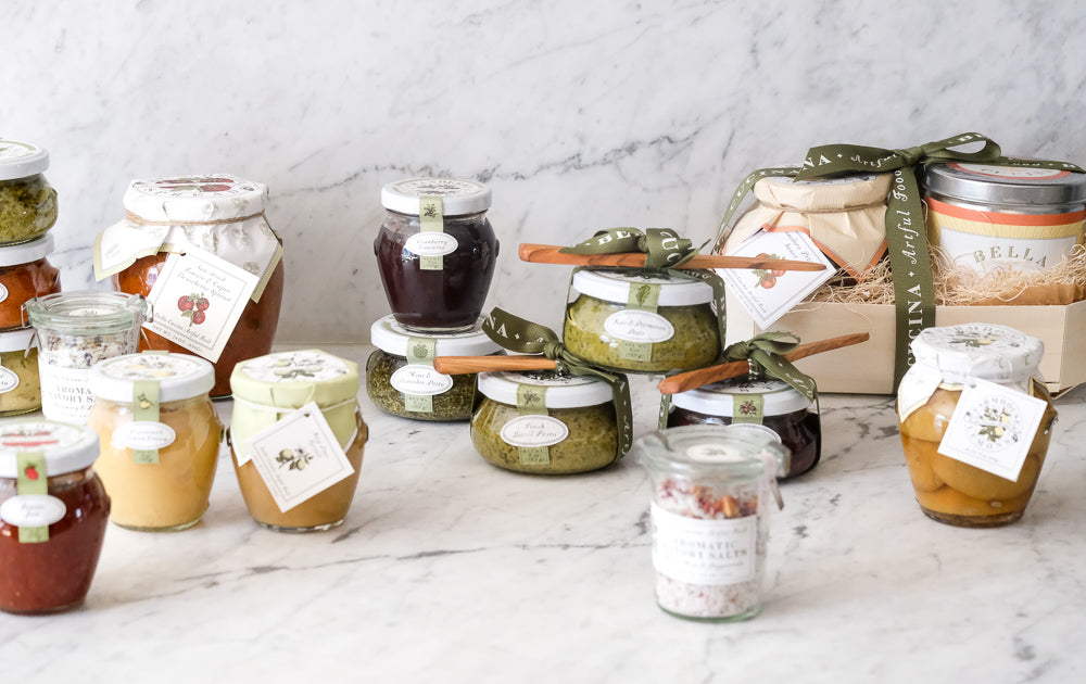 A variety of jars filled with preserves, jams, and spreads are arranged on a marble countertop. Some jars are labeled with tags, and a gift box with ribbon is included in the display.