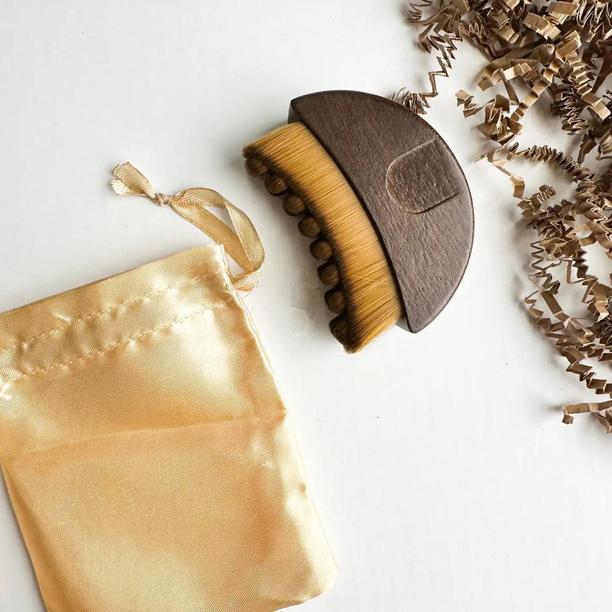 A SMILE BOUTIQUES LYMPHATIC BRUSH WITH NATURAL WOOD, featuring soft bristles, is displayed next to a satin drawstring pouch and brown paper packing material on a white surface. This elegantly designed facial massage brush is crafted from premium natural wood, making it a luxurious addition to your skincare routine.