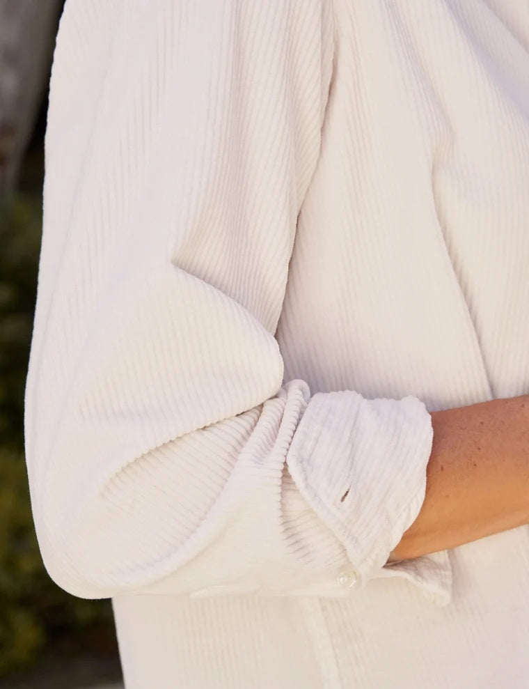 A woman stands outdoors with her hands in her pockets, looking down, wearing the FRANK &amp; EILEEN EILEEN Relaxed Button Up Shirt in Italian Corduroy Vintage White paired with blue jeans. Lush greenery and a stone building create a serene backdrop.