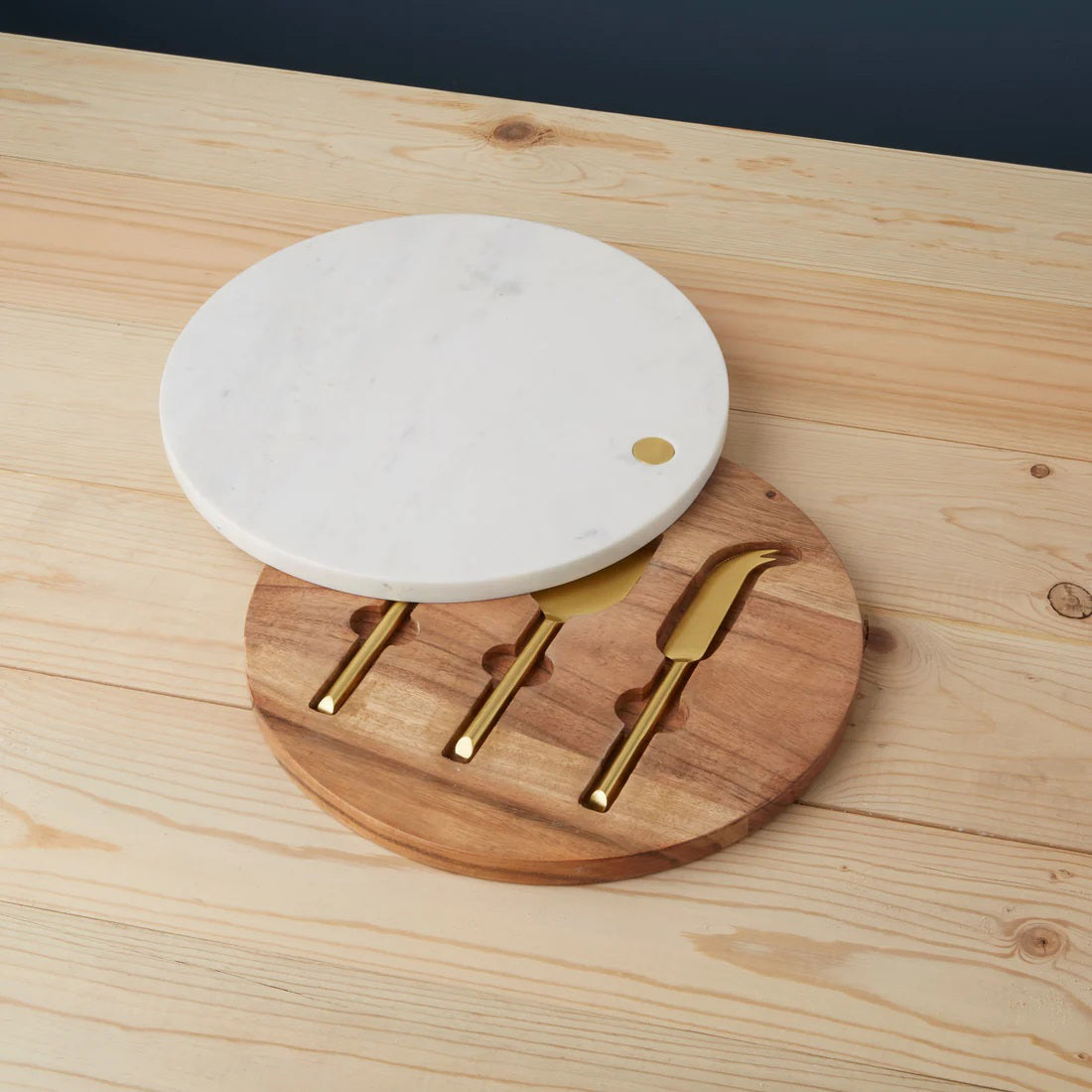 The BE HOME - GARNET Swivel Board &amp; Cheese Set by BE HOME, INC features a round white marble cheese board on a wooden base with slots for three matte gold cheese knives, offering a chic and functional addition to your table.
