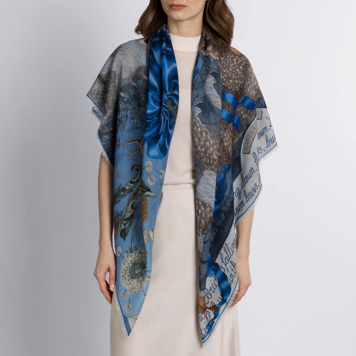 The SABINA SAVAGE ENGLAND cashmere scarf, named "London Bridge is Falling Down," displays intricate nature-themed illustrations of rabbits, flowers, and baby lambs on a light blue background. Tied in a loose knot, the scarf beautifully highlights its detailed patterns.