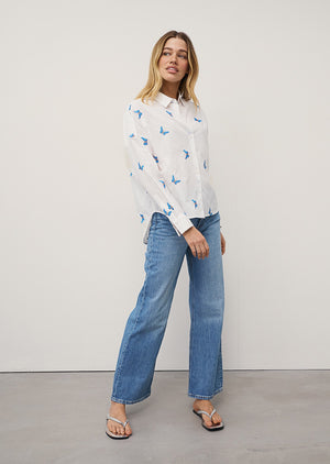 A woman with long, straight hair stands against a plain background wearing the 0039 ITALY LUCY BLOUSE WITH EMBROIDERED BUTTERFLIES, light blue wide-leg jeans, and silver sandals. She is looking slightly to the side.