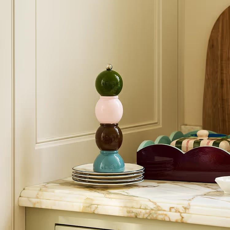 Three vibrant ADDISON ROSS LONDON Bobbin Salt or Pepper Mills (24cm) from ADDISON ROSS are stacked on a kitchen counter near wooden cabinets, with jars of dry food visible in the background.