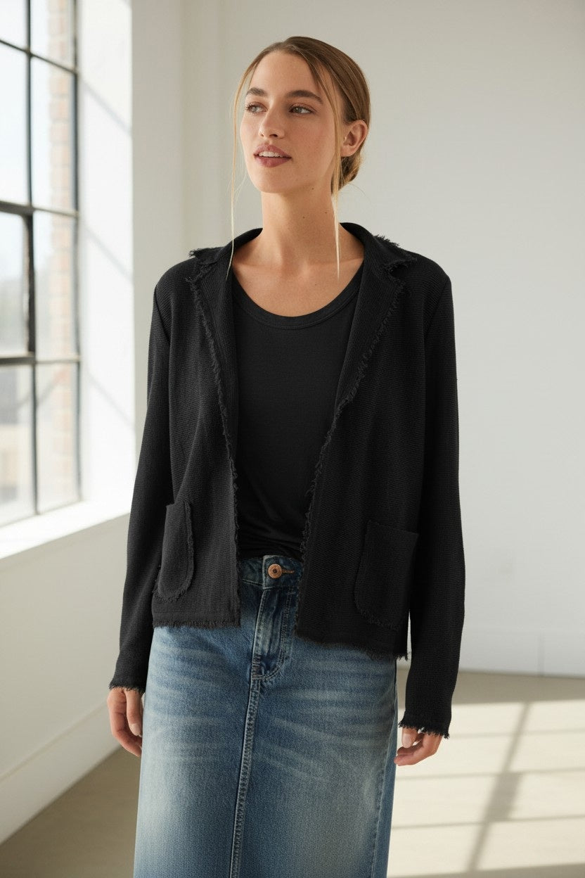 Under the soft glow of natural indoor light, a woman exudes quiet confidence as she gazes to the side, her light brown hair pulled back. She wears a black top and blue denim skirt, elevating her effortless look with a black blazer—but for versatile, lightweight layering, she could simply swap in the BASE MILANO - COTTON KNIT CARDIGAN IN BLACK by BASE MILANO.