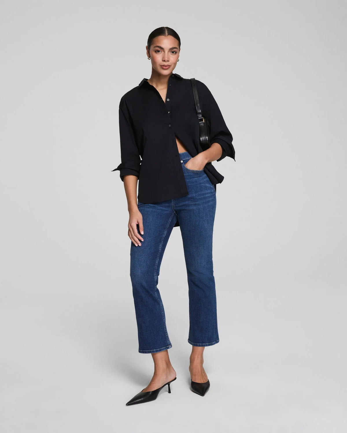 A woman with her hair pulled back stands against a plain background wearing SPANX Redefine Kick Flare Jeans in Medium Indigo, a black button-up shirt, black pointed-toe heels, and carries a black shoulder bag with one hand in her pocket.