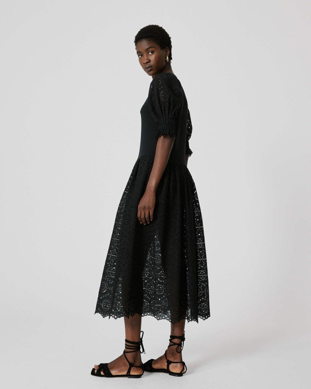 A person stands against a plain background wearing the SFIZIO - BRODERIE ANGLAISE MIDI DRESS by SFIZIO, which features elbow-length puffed sleeves and a midi-length skirt adorned with intricate lace patterns. They are also wearing black ankle-strap sandals.