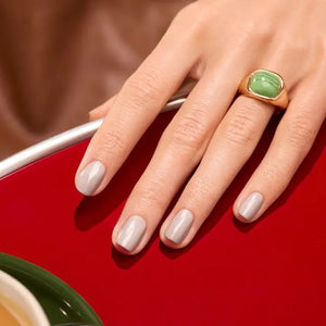 A hand painted with MANUCURIST - GREEN™ NAIL POLISH IN TWEED by MANUCURIST INC, a short pearly grey shade, rests on a shiny red surface. The person wears a gold ring featuring a large green stone on their middle finger, while a green and white bowl is partially visible in the corner.