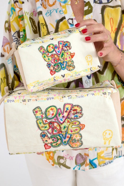 A person with painted nails holds two STORIATIPIC - LOVE COSMETIC BAG SMALL by STORIATIPIC, featuring cream cotton, colorful graffiti-style "LOVE" text and doodles, paired with a vibrant abstract-patterned shirt.
