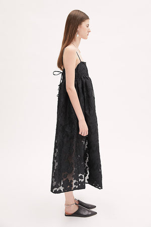 A woman wears the MEIMEIJ Lace Empire Waist Dress, a sleeveless black floral lace midi with spaghetti straps and a sheer overlay. The flowing fit makes it perfect for a garden party. She stands against a plain white background.
