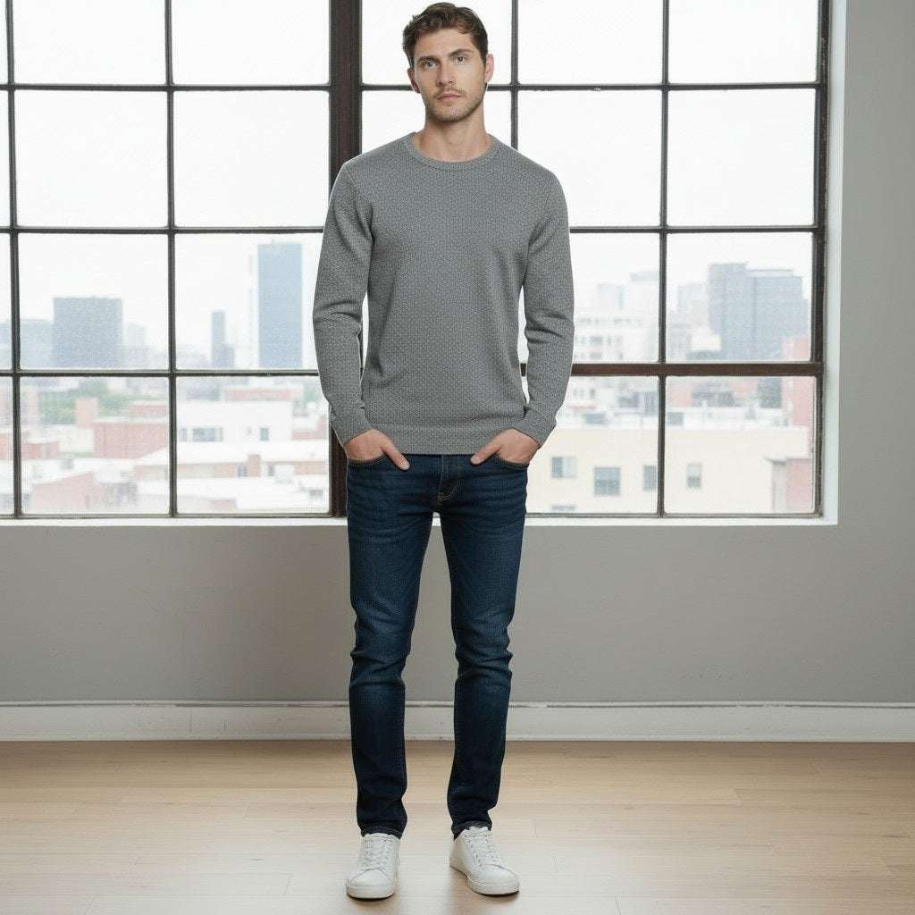 A man stands indoors in front of large windows, wearing the AUTUMN CASHMERE - CASHMERE BASIC CREW NECK WITH RIB BOTTOM IN STONE, paired with dark blue jeans and white sneakers. The cityscape visible outside the windows complements his refined taste in men’s knitwear from AUTUMN CASHMERE.