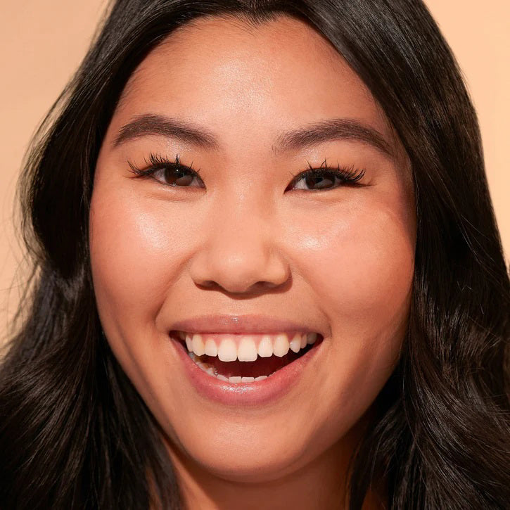 A close-up of a person with natural curly hair, adorned in gold hoop earrings and a white top. They wear a neutral facial expression while looking directly at the camera. The softly lit, plain background enhances their glowing skin as if touched by JASON WU - TINTED MOISTURIZER MEETS CC CREAM from JASON WU BEAUTY.