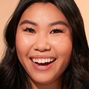 A close-up of a person with natural curly hair, adorned in gold hoop earrings and a white top. They wear a neutral facial expression while looking directly at the camera. The softly lit, plain background enhances their glowing skin as if touched by JASON WU - TINTED MOISTURIZER MEETS CC CREAM from JASON WU BEAUTY.