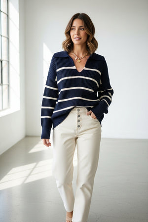 Wearing the MERSEA Camden Sailor Sweater in Navy Cream—adorned with classic white stripes—a woman stands in a sunlit room with expansive windows, smiling as she pairs the look with cream high-waisted pants, effortlessly channeling coastal Maine charm.