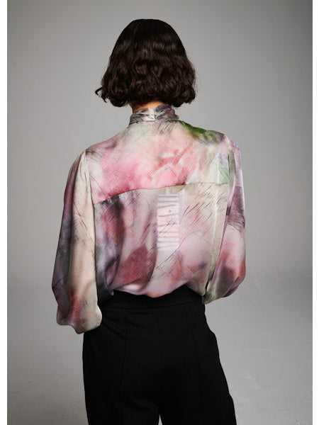 A woman with short dark hair wears the MILVA MI - BRUSHSTROKE BLOUSE by MILVA, a pastel long-sleeved statement bow top, paired with high-waisted black pants. She poses with eyes closed against a plain light background.