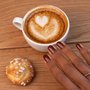 A hand adorned with MANUCURIST - GREEN™ Nail Polish in Chestnut by MANUCURIST INC rests on a wooden table beside a cup of coffee with heart-shaped latte art and a small pastry sprinkled with coarse sugar.