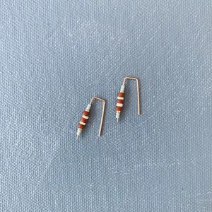 ALICE RISE - LITTLE QUILL EARRINGS IN TERRACOTTA by ALICE RISE rest on a textured gray surface, their unique design reminiscent of bent electronic resistors—perfect inspiration for handmade jewelry enthusiasts.