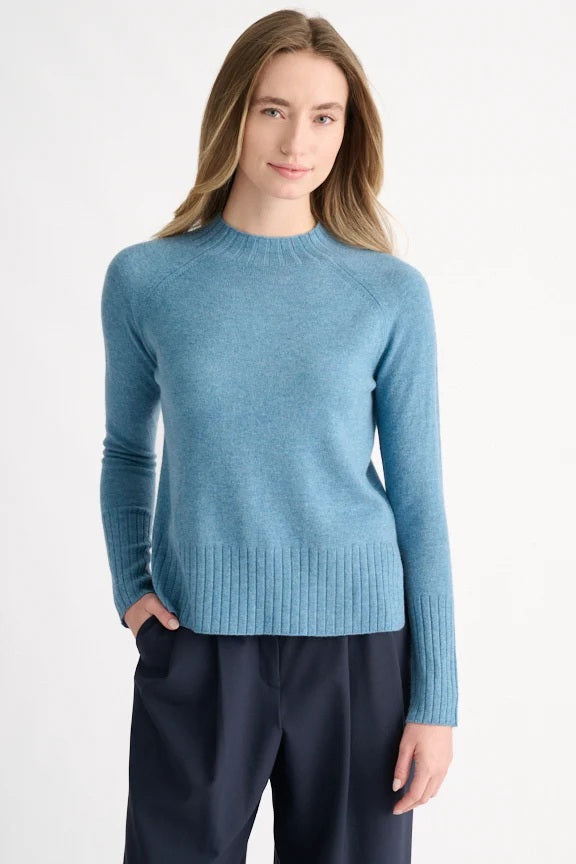 A woman with blonde hair models the KINROSS CASHMERE Hi Low Rib Trim Funnel in Loch—a light blue, long-sleeve cashmere pullover with ribbed trim—paired with dark pants. She stands against a plain white background, turned slightly to the side.