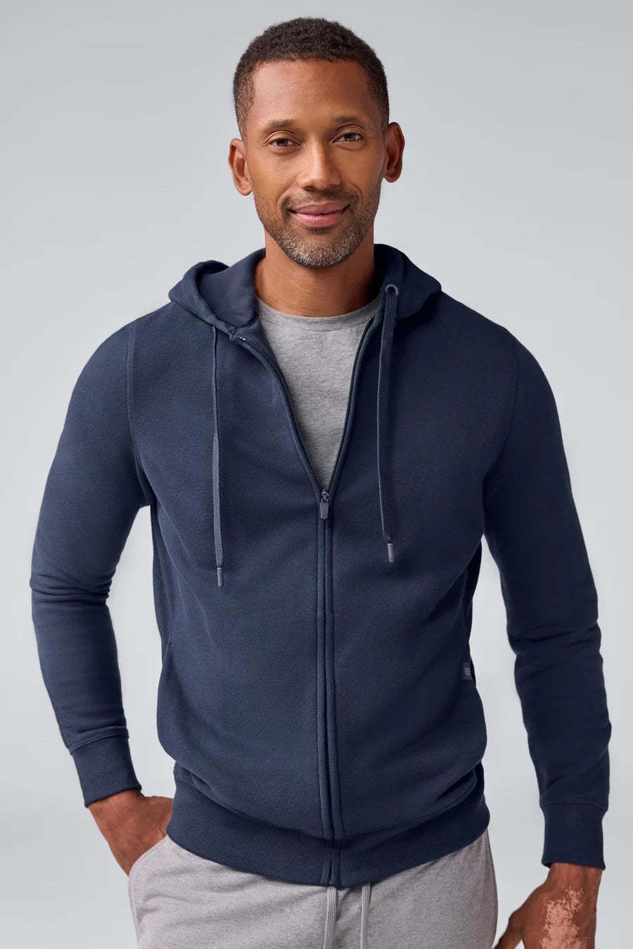 A man wears the MACK WELDON Ace Full-Zip Sweatshirt in True Navy (Bird's Eye) with light gray pants, standing against a plain light gray background and looking slightly to his left with a neutral expression.