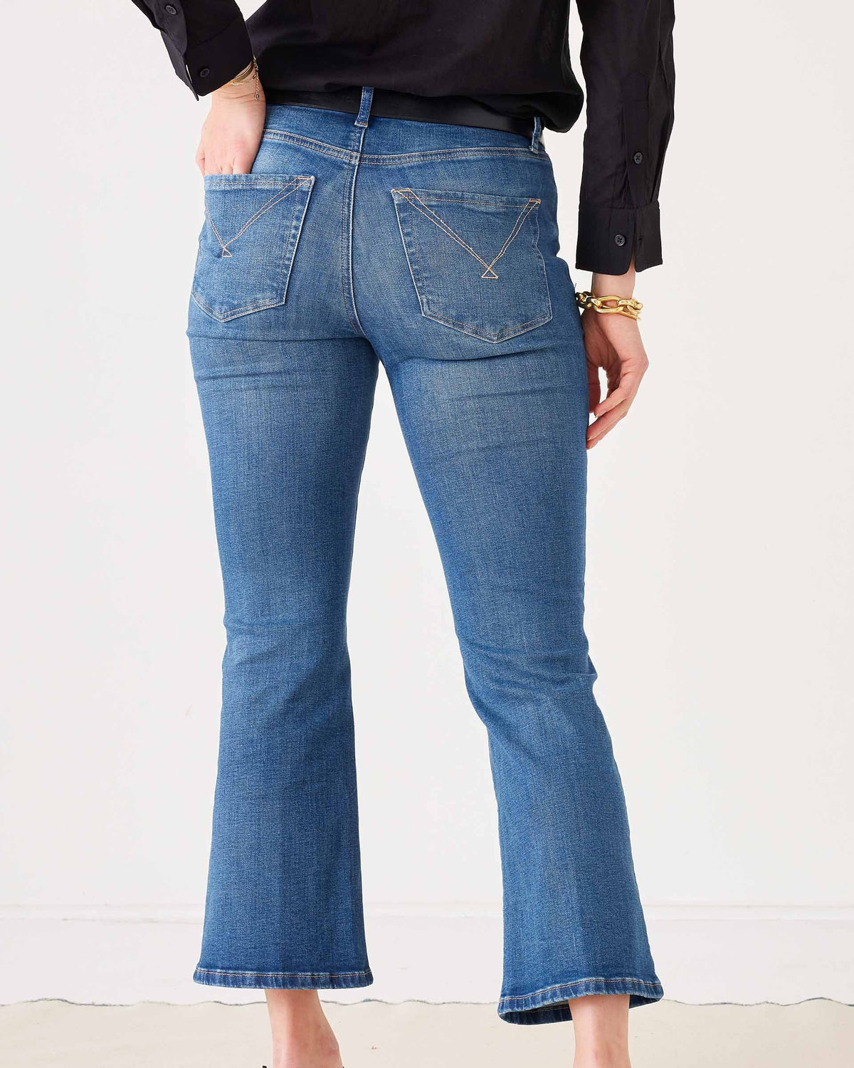 A woman stands against a plain white background wearing sunglasses, a black long-sleeve blouse, black heels, and holding a black handbag. She completes her look with the MERSEA Murphy Mini Boot Jean in Tide from MERSEA—blue, cropped organic cotton jeans designed for a gap-free waist.