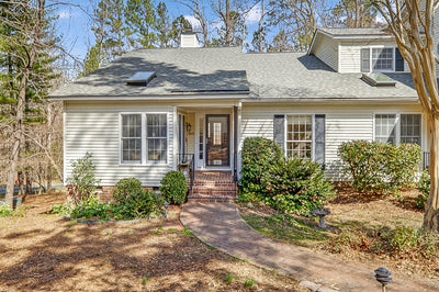 A light-colored house with a gray roof, front porch, brick steps, and a curved brick walkway. The yard has shrubs and trees, with forested areas visible in the background under a blue sky.