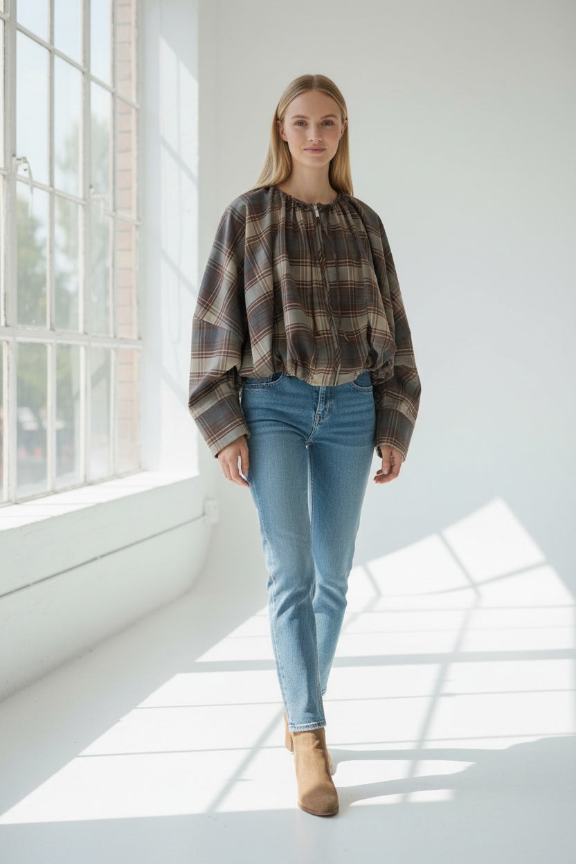 A woman stands in a bright room, wearing the MEIMEIJ plaid jacket with sculptural volume, paired with blue jeans and tan ankle boots. Sunlight streams through large windows, casting shadows on the floor.