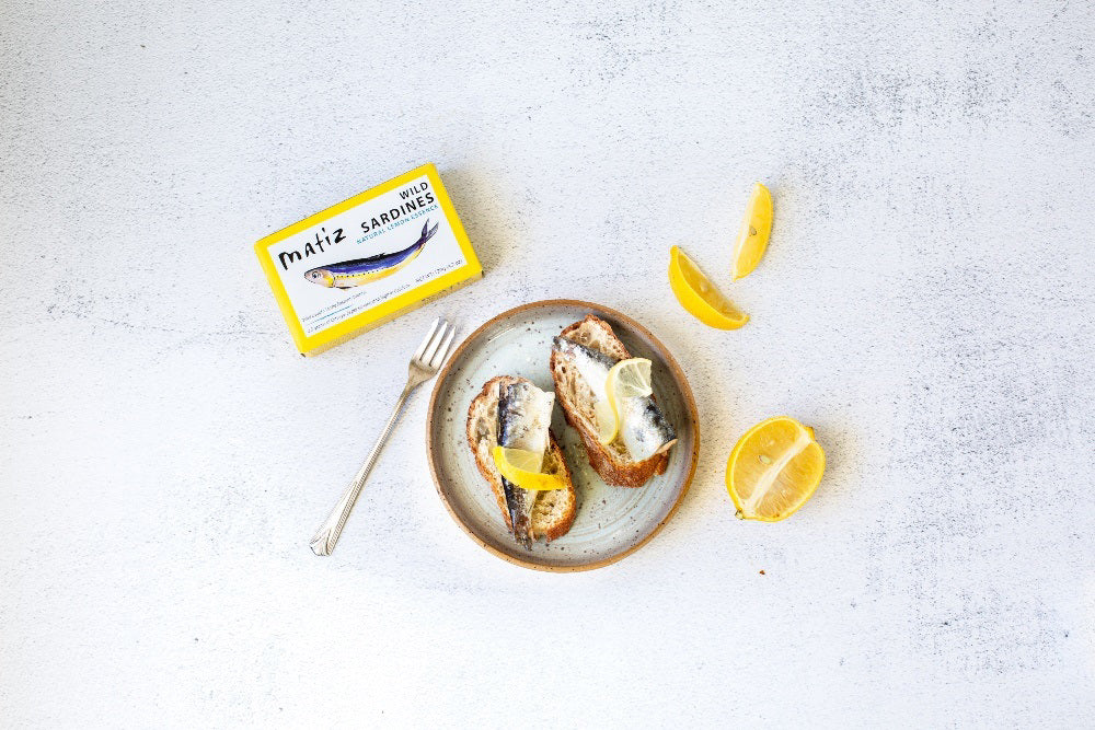 MATIZ ESPANA's MATIZ - SARDINES WITH LEMON comes in a yellow box featuring a sardine illustration. Key highlights include "Wild caught in the Eastern Atlantic," "2.3 grams of Omega-3s," "High in Calcium," and "Rich flavor." Contains 120g (4.2 oz); includes Spanish olive oil and lemon essence.