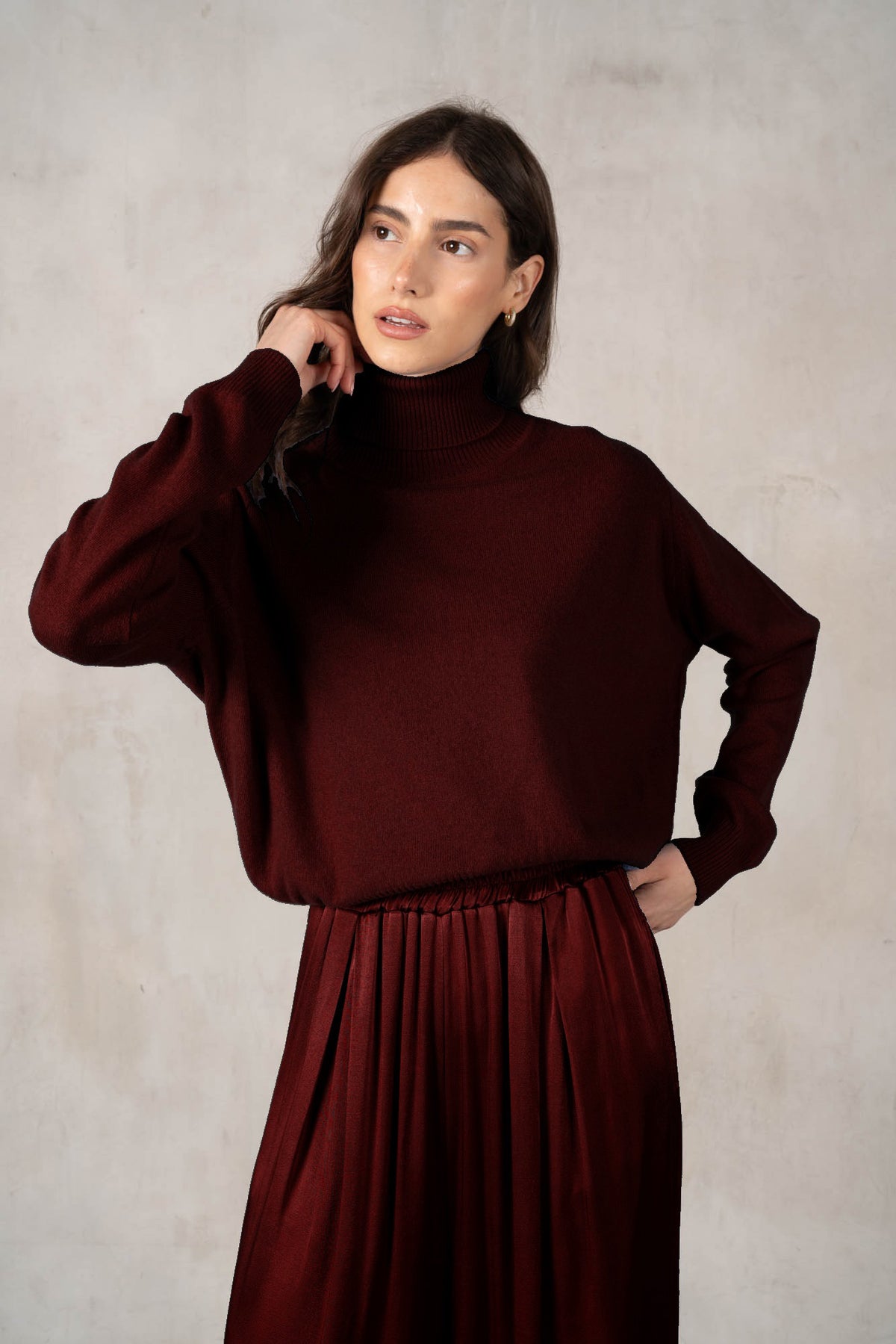 A woman with long brown hair smiles against a white background, wearing a ROSE &amp; CROWN - CASHMERE TURTLENECK and wide-legged green pants, accessorized with necklaces, bracelets, and a large black handbag.