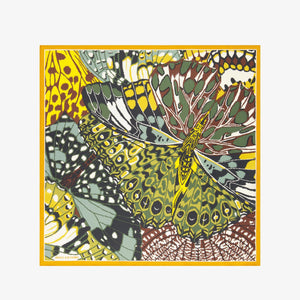 A woman with dark hair wears a beige blazer and the INOUI EDITIONS - PAPILLON (BUTTERFLIES) Silk Scarf in Green by INOUI, a versatile accessory made in India, featuring vibrant yellow, black, and green designs, against a plain white background.