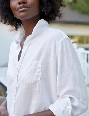 A person with curly hair wears the FRANK & EILEEN EILEEN Relaxed Button Up in Washed Linen White, standing outdoors against a blurred background of greenery and a building.