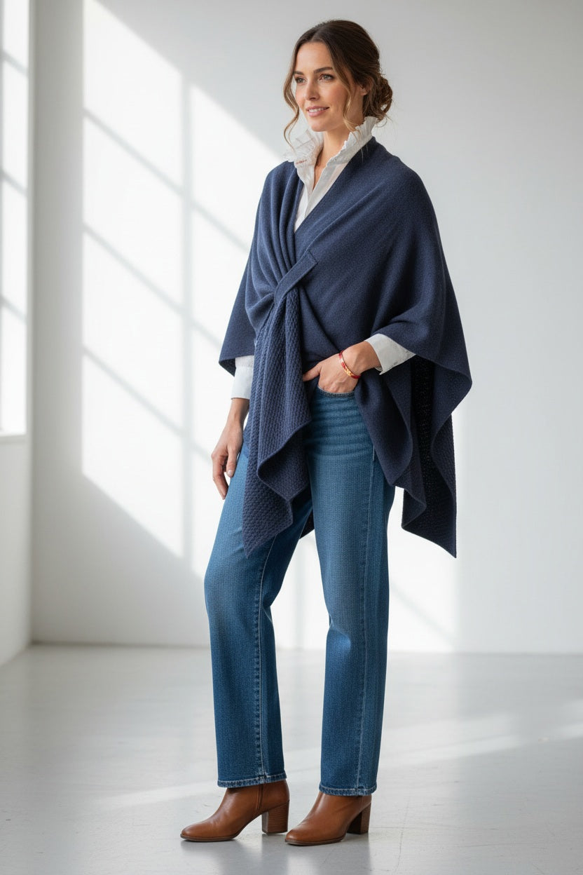 A woman stands indoors wearing the MERSEA - Modern Travel Wrap in Slate draped over a light gray patterned shirt, beige wide-leg pants, and tan shoes. She looks at the camera with a relaxed expression and long blonde hair.
