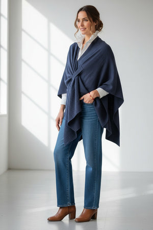 A woman stands indoors wearing the MERSEA - Modern Travel Wrap in Slate draped over a light gray patterned shirt, beige wide-leg pants, and tan shoes. She looks at the camera with a relaxed expression and long blonde hair.