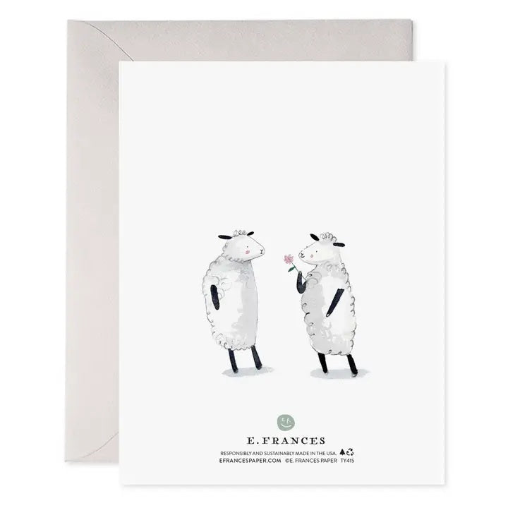 E. FRANCES - THANK EWE CARD by E. FRANCES PAPER features a watercolor design with two cartoon sheep, one holding a pink flower behind its back. The cheerful words "THANK EWE" appear above them in playful lettering. A gray envelope is partially visible behind the card. Proudly made in the USA.