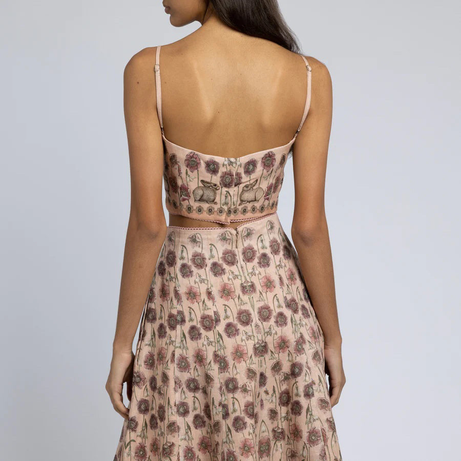 A woman is wearing a light pink floral two-piece outfit. The SABINA SAVAGE - HONOURING ARGOS CROPPED FITTED TOP from SABINA SAVAGE ENGLAND is sleeveless with thin straps and a square neckline, paired with a high-waisted skirt that flows gracefully from the hips. Crafted from pure linen, the fabric features a pattern of small, detailed floral designs perfect for any Greek-inspired look.