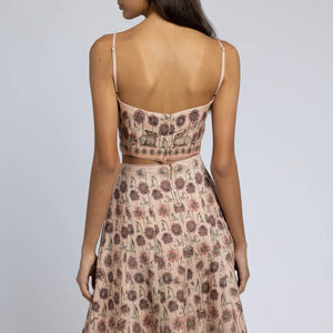 A woman is wearing a light pink floral two-piece outfit. The SABINA SAVAGE - HONOURING ARGOS CROPPED FITTED TOP from SABINA SAVAGE ENGLAND is sleeveless with thin straps and a square neckline, paired with a high-waisted skirt that flows gracefully from the hips. Crafted from pure linen, the fabric features a pattern of small, detailed floral designs perfect for any Greek-inspired look.