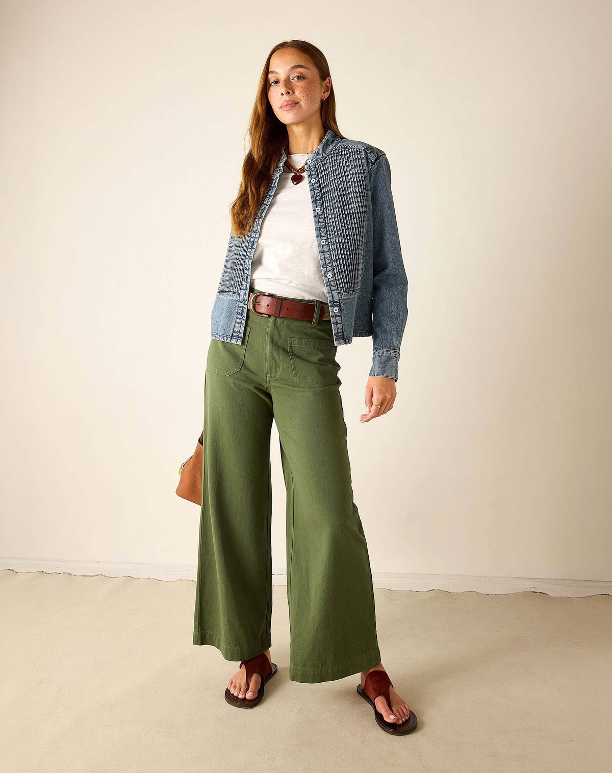 A woman with long brown hair and a relaxed expression stands in a neutral room wearing a denim jacket, white shirt, brown belt, sandals, and holding a tan bag. She pairs her outfit with MERSEA’s Londonberry Twill Pant in Olive for a laid-back look.