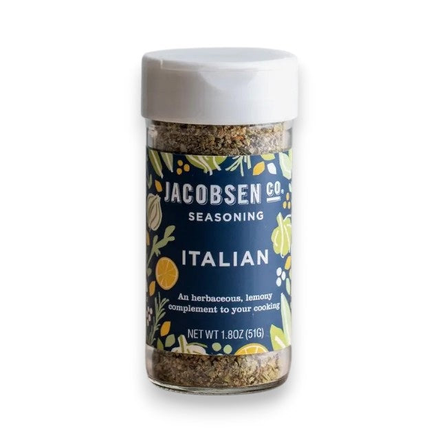 A jar of JACOBSEN SALT - ITALIAN SEASONING from JACOBSEN SALT CO. sits on a kitchen counter near utensils, a mason jar with a metal lid, and a ceramic cup. The spice jar label features a blue and yellow floral design.