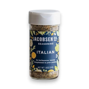 A jar of JACOBSEN SALT - ITALIAN SEASONING from JACOBSEN SALT CO. sits on a kitchen counter near utensils, a mason jar with a metal lid, and a ceramic cup. The spice jar label features a blue and yellow floral design.