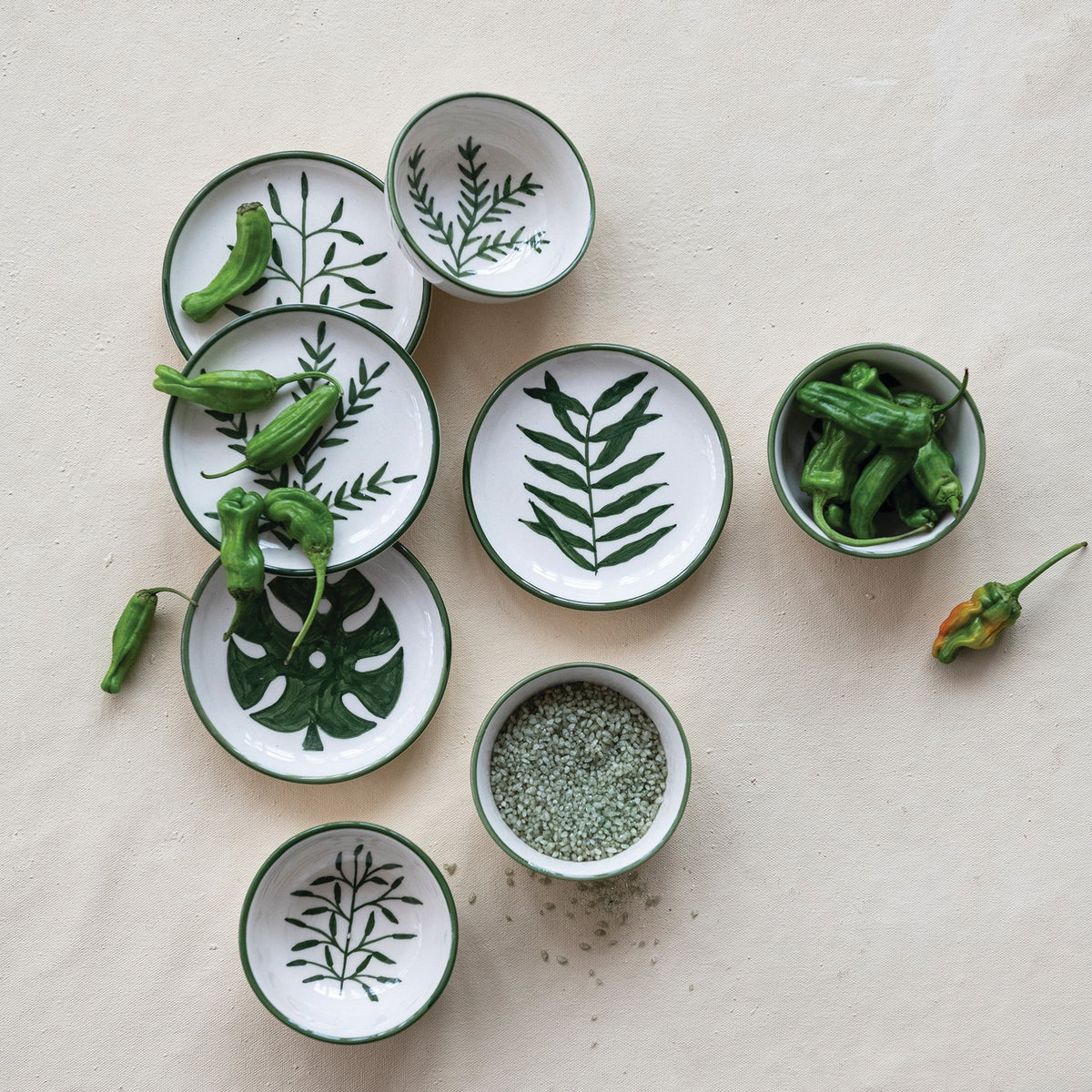 CREATIVE COOP's HAND-PAINTED STONEWARE BOWLS WITH BOTANICAL DESIGN, some holding green peppers or grains, are arranged on a light beige surface—each bowl highlights its unique handcrafted artistry.