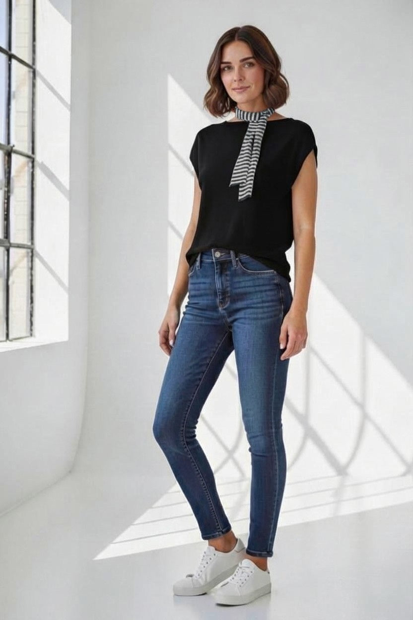 A woman stands in a bright room wearing the FLOOR Cotton Top with Stripe Neck Tie, paired with blue skinny jeans and white sneakers. Sunlight streams through large windows, casting shadows on the white walls and floor.
