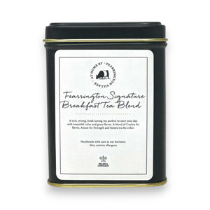 The Fearrington Signature Collection's black tin container features a white label with the text "Fearrington Signature Breakfast Tea Blend" from Fearrington House. The label describes this loose leaf tea as a rich, robust blend of Ceylon and Assam. The bottom of the tin bears a logo and text indicating that it is handmade.