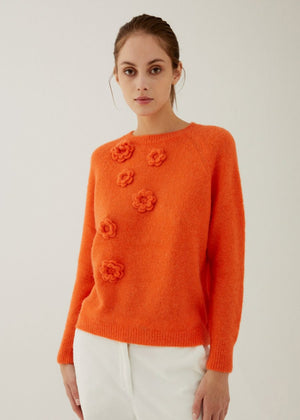 A woman with light brown hair wears the ANNECLAIRE hand embroidered crew neck wool sweater in light green with flower details and a white skirt, standing against a pale background. The timeless piece is handmade in Italy.