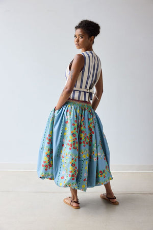A person stands indoors wearing a white and blue striped vest and the HOPE FOR FLOWERS Bubble Hem Skirt in Chambray Floral by HOPE FOR FLOWERS. They have short curly hair and are wearing sandals.