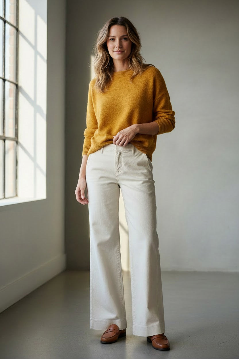 A person stands on grass with hands on hips, wearing FRANK &amp; EILEEN's West Village NYC Wide Leg Trouser in Italian Performance Linen Cement and a white top, facing away from the camera with a hedge and trees in the background.