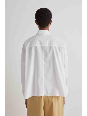 A person wears the MEIMEIJ - COTTON RUFFLE BLOUSE, featuring a prominent front ruffle, paired with wide light brown pants, standing against a plain light background.