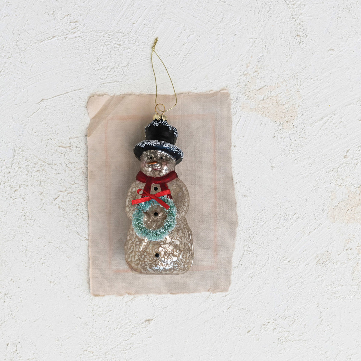 The CREATIVE COOP MERCURY GLASS SNOWMAN ORNAMENT features a vintage look with a black top hat, red scarf, green wreath, gold string hanger, and an antique-style finish.